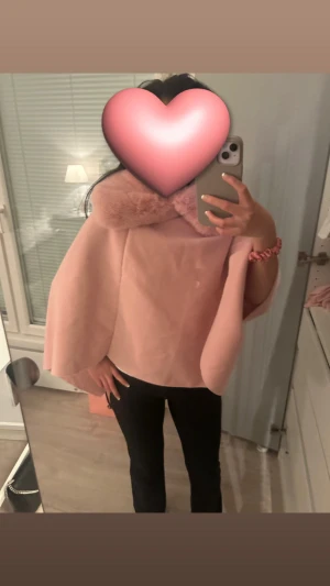 Ljusrosa kappa/poncho i kappmaterial - Säljer en ljusrosa kappa/poncho med bred krage i fluffig pälsimitation. Jackan har en elegant och loose passform med vida ärmar som ger en cool och trendig look. Perfekt för dig som vill sticka ut med en mjuk och feminin vibe.