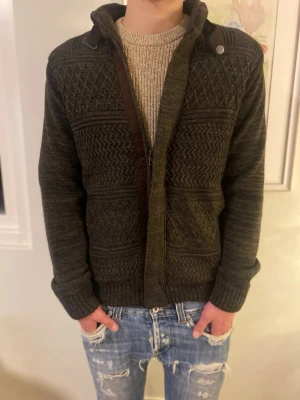 Mörbrun stickad kofta/jacka  - Säljer en mörkbrun stickad kofta med quiltat foder. Koftan har dragkedja, ribbade muddar och snygga mönster i stickningen. Perfekt för kyliga dagar och har hög krage med knappdetalj. Storlek är M men är lite stor i storleken, Killen på bilder är 182 och väger 70kg.