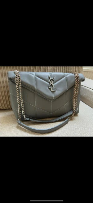 Grå quiltad axelväska  - Snygg grå axelväska med quiltad design och silverfärgad kedja. Väskan har ett stort emblem i metall på framsidan och praktiskt axelband i skinn. Perfekt för dig som gillar stilrena detaljer och exklusiv känsla.Köparen betalar frakten