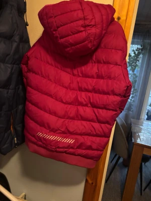 Mörkröd pufferjacka med huva - Säljer en mörkröd pufferjacka med huva och dragkedja framtill. Jackan har två fickor med dragkedja och ribbad nederkant. Materialet är syntet och jackan är quiltad för extra värme. Perfekt för kalla dagar och har en snygg, sportig look.