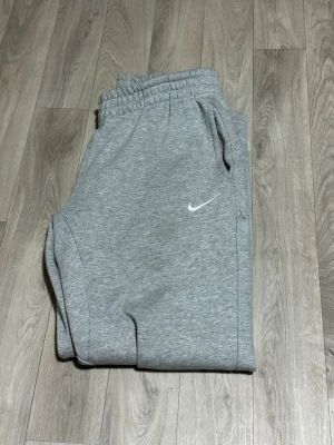 Grå mjukisbyxor från Nike, storlek L - Säljer ett par grå mjukisbyxor från Nike i storlek L. Byxorna har elastisk midja med snörning, två sidofickor och en vit Nike-logga på benet. Materialet är mjukt och skönt, perfekt för chill eller träning. Klassisk loose passform.