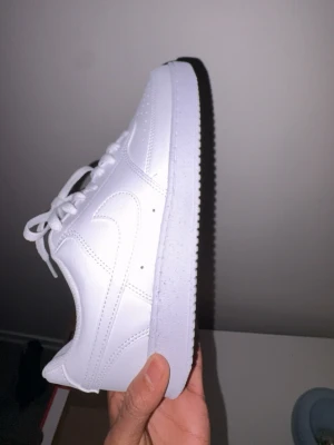 Nike Court Vision Low vit sneaker - Nike Court Vision Low sneakers i helvit färg med klassisk snörning och rund tå. Snyggt och cleant utseende med perforerade detaljer på tån och sidopaneler. Tillverkade i syntetmaterial och skinn, med platt sula och låg profil. Perfekt för dig som gillar stilrena sneakers.