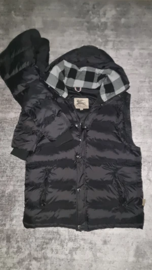 Svart jacka från Burberry XXL - Säljer en svart dunväst från Burberry i storlek XXL. Västen har en quiltad look, avtagbara armar, luva med grått och svart rutigt foder, samt snygga metallknappar med Burberry-gravyr. Två dragkedjefickor framtill och klassisk Burberry-label på insidan. Aldrig använt. Möjlighet för byten finns alltid