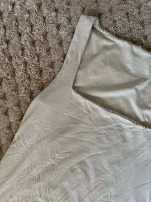 Beige ärmlös linnetopp med fyrkantig hals - Säljer en stilren beige linnetopp med fyrkantig halsringning. Toppen är ärmlös och har en slät, mjuk yta i syntetmaterial som ger en lätt glans. Perfekt för varma dagar och enkel att matcha med olika outfits.