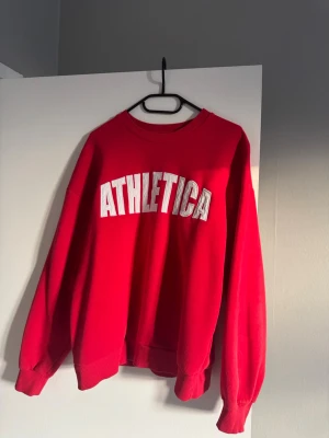 Röd sweatshirt från Gina Tricot - Cool röd sweatshirt från Gina Tricot med ATHLETICA-tryck i vitt på bröstet. Tröjan har rund hals, ribbade muddar och lång ärm. Perfekt för en sportig och avslappnad stil. Mjuk insida och loose passform.