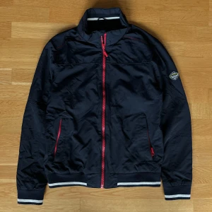 Mörkblå bomberjacka från Erla of Sweden - Snygg mörkblå bomberjacka från Erla of Sweden med röda detaljer och dragkedjor. Jackan har ribbade muddar med vita ränder, ståkrage och ett coolt märke på ärmen. Materialet är polyester och jackan har två fickor framtill. Armlängd: 61cm. Längd: 65cm Bredd: 43cm. Hör av dig vid frågor pris kan diskuteras