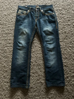True Religion Rocco jeans, blå, stl 34 - Säljer ett par blå True Religion Rocco jeans med kontrasterande grova sömmar och ikoniska broderade fickor bak. Jeansen har klassisk femficksdesign, låg midja och raka ben. Materialet är slitstarkt denim i bomull med coola detaljer i orange och beige.
