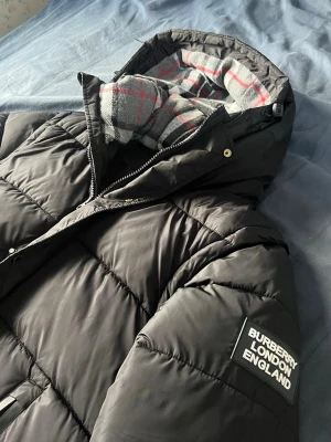 Svart pufferjacka från Burberry - Svart pufferjacka från Burberry med stor huva och rutig grå, vit och röd insida. Jackan har två dragkedjeförsedda fickor framtill och en tydlig Burberry London England-patch på ärmen. Quiltad design och klassisk pufferstil, perfekt för kalla dagar.