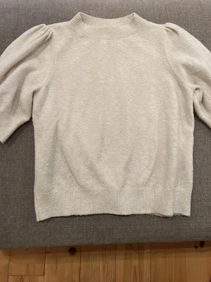 Beige stickad tröja från Vero Moda - Supersöt beige stickad tröja från Vero Moda med puffärmar och ribbade muddar vid hals, ärmslut och nederkant. Perfekt för en chill men ändå snygg look. Tröjan har en mjuk känsla och en enkel, stilren design.
