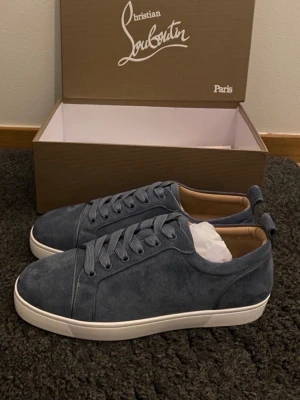 Blå mocka sneakers från Louboutin - Snygga blå sneakers från Christian Louboutin i mjuk mocka med klassisk vit sula och ikonisk röd undersida. Skorna har rund tå, snörning och beige innersida i skinn. Perfekta för dig som vill ha något exklusivt och stilrent.