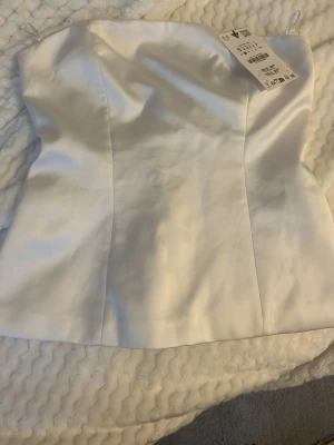 Vit satin bandeau tubtopp S - Säljer en stilren vit bandeau tubtopp från Zara i storlek S. Toppen är axelbandslös och har en glansig satinfinish som ger en lyxig känsla. Den är aldrig använt då den inte passar. 