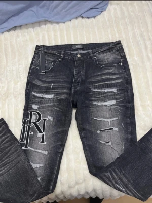 Svarta jeans från Amiri med tryck - Säljer ett par svarta jeans från Amiri i storlek 34. Jeansen har slitna detaljer och ett stort vitt tryck med bokstäver på ena benet. De har fem fickor, knapp och dragkedja i midjan samt en cool tvättad look. Perfekt för dig som gillar streetwear och statement-plagg.