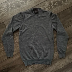 Grå stickad tröja från Oscar Jacobson - Säljer en stilren grå stickad tröja från Oscar Jacobson i storlek S. Tröjan har rund hals, ribbade muddar vid ärmslut och nederkant samt långärmad passform. Perfekt för dig som gillar klassisk och enkel design.