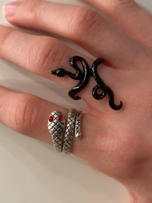 Två ringar med ormmotiv - Säljer två coola ringar med ormmotiv. En är i silverfärgad metall med skalmönster och röda ögon, den andra är svart och formad som en slingrande orm. Perfekta accessoarer för dig som gillar edgy och unika smycken. Ej äkta silver! Styckpris 10kr, båda för 15kr 🫶