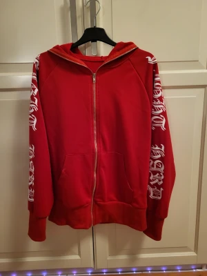 Röd zip up hoodie med vit text på ärmar  - Säljer en röd zip up hoodie. Den är lite tunn men är väldigt skön. 