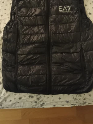 Svart dunväst EA7 Emporio Armani - Säljer en svart dunväst från EA7 Emporio Armani med quiltad design och dragkedja framtill. Västen har en stilren look med logotypen tryckt på bröstet. Perfekt för lager-på-lager och passar dig som gillar exklusiva märken.