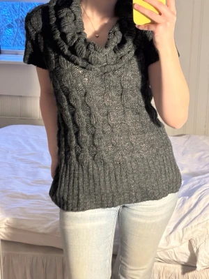 Mörkgrå stickad polotröja - Säljer en mörkgrå stickad polotröja med korta ärmar och grov kabelstickning. Tröjan har en bred, rullad polokrage och ribbade detaljer vid nederkant och ärmslut. Perfekt för lager-på-lager under kyliga dagar.