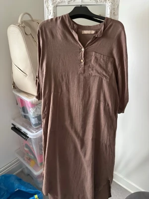 Brun linnéklänning från Marta - Säljer en brun långklänning från Marta i mjukt linne. Klänningen har trekvartsärm, knappar framtill, en bröstficka och två sidofickor. Enkel och chill design med liten krage och slits nedtill. Perfekt för dig som gillar avslappnad stil.
