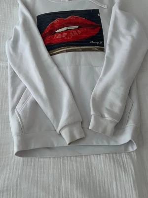 Hoddie med pussmun the cool elephant  - En väldigt fin och mysig hoddie från the cool elephant. Hoodien har en fin och rymlig ficka på magen och den har även ett fint tryck med en pussmun på magen. Denna hoodie är perfekt för alla tillfällen, mysiga kvällar, umgås med vänner osv. Hoddien är i storlek XS men den är stor i storleken 