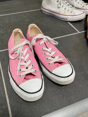 Rosa Converse All Star sneakers - Säljer ett par klassiska Converse All Star sneakers i rosa med vit tå och vit sula. Skorna har snörning med vita skosnören och metallhål, samt en rund tå och platt sula. Materialet är bomullstyg och de har den ikoniska svarta linjen runt sulan.