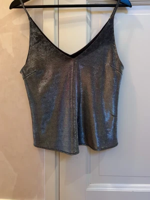 Glittrig silver linnetopp från Stay - Säljer en snygg glittrig silverfärgad linnetopp från Stay i storlek M. Toppen har smala axelband och v-ringad hals, perfekt för att sticka ut. Materialet är syntet med en skimrande finish som fångar ljuset. Passformen är normal och toppen är ärmlös.