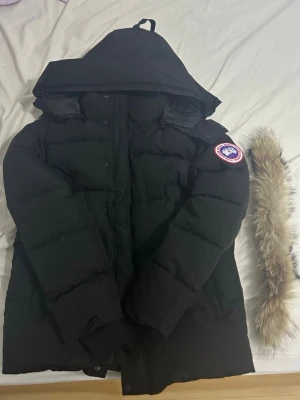 Svart pufferjacka från Canada Goose - Säljer en svart pufferjacka från Canada Goose med stor huva och avtagbar päls. Jackan har klassisk logga på ärmen, dragkedja och knappar framtill. Perfekt för kalla vinterdagar och har en riktigt schysst vinter look. Det är storlek S men den är lite oversized så den passar även M