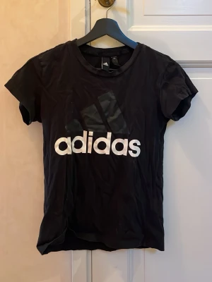 Svart Adidas t-shirt med logga - Svart t-shirt från Adidas i storlek XS med stor vit Adidas-logga på bröstet. Klassisk rund hals och korta ärmar. Tillverkad i mjuk bomull som är skön mot huden. Perfekt för dig som gillar sportig och enkel stil.