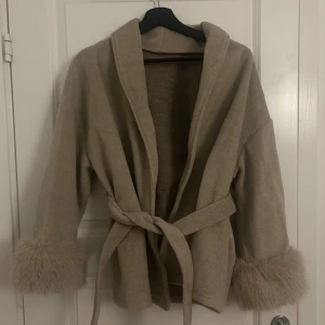 Beige omlott kappa med fuskpäls - Snygg beige omlott kappa med bälte i midjan och fluffiga fuskpälsdetaljer vid ärmsluten. Jackan har en loose passform och är tillverkad i mjukt material som känns lyxigt. Perfekt för dig som vill ha en chill och trendig look. Köpte den för 800kr i Lamuor modest. Har alldrig haft på mig den.