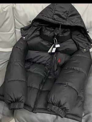 Svart pufferjacka från Polo Ralph Lauren - Säljer en svart pufferjacka från Polo Ralph Lauren med huva och dragkedja. Jackan har en röd broderad logga på bröstet, elastiska muddar och stora puffiga sektioner som håller dig varm. Perfekt för kalla dagar och riktigt snygg streetstil.