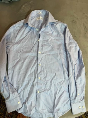 Ljusblå skjorta från These Glory Days - Snygg ljusblå skjorta från These Glory Days med klassisk krage och vita knappar. Skjortan har lång ärm och är tillverkad i mjuk bomull. Perfekt för dig som gillar stilren och enkel design med skandinavisk touch.du behöver bara stryka skjortan efter blir det som ny annars så kan du returnera direkt. 