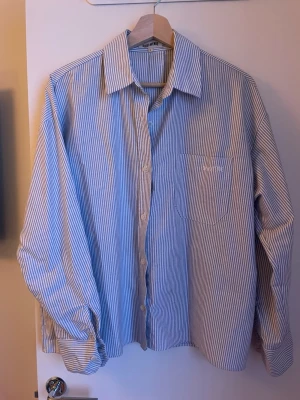 Blåvit randig skjorta från Djerf Avenue - Säljer en blåvit skjorta från Djerf Avenue i storlek S. Skjortan har klassisk krage, bröstficka med broderad logga och knappar framtill. Den är långärmad och har ett snyggt randigt mönster som ger en fräsch vibe. Perfekt för dig som gillar stilrena plagg. 