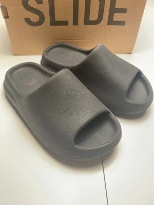 Adidas Yeezy Slides  - Säljer riktigt fina svarta Adidas Yeezy Slides. De har en riktigt bekväm passform och är riktigt snygga. Går att styla till väldigt mycket och är perfekta inför sommaren. Sulans innermått är 24 cm. De är i princip oanvända. Säljer på grund av att de inte passar mig. Priset är inte hugget i sten!