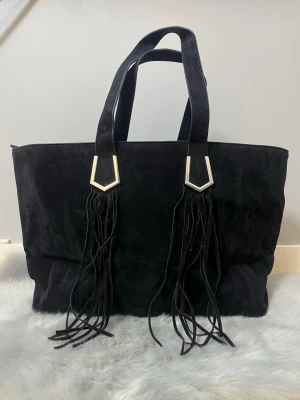 Svart väska med fransar från Bershka polen  - Stor svart väska i mocka med två rejäla handtag och snygga fransdetaljer som hänger från metallbeslag. Väskan har en enkel, boxig form och är perfekt för dig som gillar att bära med dig mycket. Materialet är mjukt och ger en cool vibe.
