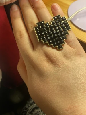Svart pixelhjärta ring - Cool ring i form av ett pixelhjärta med svarta fyrkantiga stenar och guldfärgad kant. Ringen har en chunky och unik design som verkligen sticker ut. Perfekt för dig som gillar statement-smycken och vill ha något som syns.