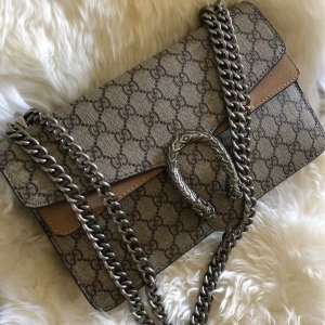 Väska med kedja och mönster - Säljer en snygg axelväska med klassiskt mönster i beige och brunt. Väskan har en robust silverfärgad kedja och ett detaljerat spänne framtill. Insidan är klädd i mocka och har ett fack med dragkedja. Perfekt för dig som gillar lyxiga accessoarer.