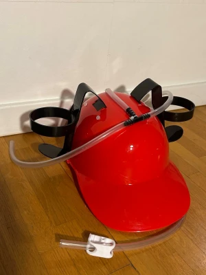 Röd öl-hjälm med sugrör - Säljer en röd öl-hjälm med två hållare för burkar och genomskinliga sugrör som går ner till munnen. Perfekt för att ha händerna fria på festen eller till roliga aktiviteter. Justerbar passform med svarta plastdetaljer.