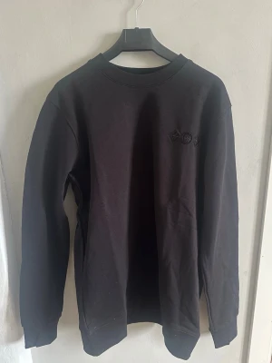 Svart sweatshirt, LV - Svart sweatshirt från L V  med broderad L V logga i stor stil på ryggen och diskret broderad detalj på bröstet. Tröjan har rund hals, långa ärmar och ribbade muddar. Materialet är mjuk bomull som känns skönt mot huden. Perfekt för dig som gillar exklusiva streetwear-vibbar.