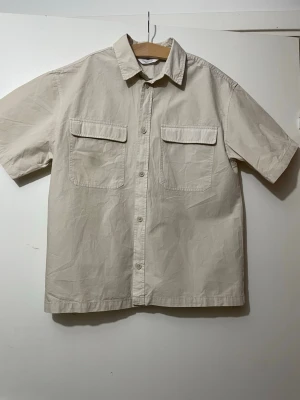 Beige overshirt skjorta kortärmad  - Snygg beige overshirt med korta ärmar och två stora bröstfickor med lock. Skjortan har klassisk krage och knäppning framtill med matchande knappar. Tillverkad i mjuk bomull som känns chill och enkel att styla till olika outfits.