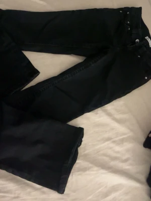 Svarta bootcut jeans med fickdetaljer - Snygga svarta jeans med bootcut passform och klassiska fem fickor. Baktill finns två fickor med lock och silverfärgade knappar. Jeansen har midjehöjd och är gjorda i ett jeansmaterial som känns slitstarkt och bekvämt. Perfekt för dig som gillar en lite vidare siluett.