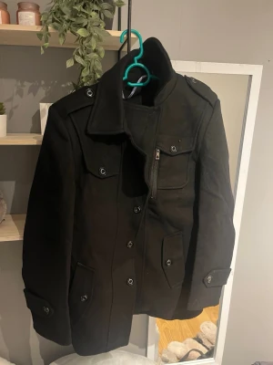 Svart rock med knappar och fickor - Snygg svart rock med hög krage, flera fickor med lock och knappar samt en bröstficka med dragkedja. Jackan har långa ärmar och dekorativa axelklaffar. Perfekt för dig som gillar stilrena och klassiska plagg. Den är liten i storleken.