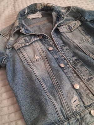 Blå jeansjacka från Calvin Klein Jeans - Snygg blå jeansjacka från Calvin Klein Jeans med slitna detaljer och klassiska silverfärgade knappar. Jackan har två bröstfickor med lock och en cool, lite tvättad look. Perfekt att slänga över en hoodie eller t-shirt för en chill vibe. Köpt för 1499 kr
