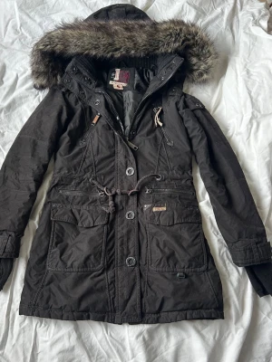 Vintage Jacka  - Svart parkajacka med stor huva och fluffig fuskpäls. Jackan har flera fickor med dragkedjor och knappar, samt snörning i midjan för snygg passform. Bra skick och jättevarm så passar jättebra nu till vintern. 