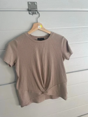 Beige ribbad topp med knut från XOXO Vibes - Säljer en beige ribbad topp från XOXO Vibes i mjukt material. Toppen har korta ärmar, rund hals och en snygg knutdetalj framtill som ger en cool twist. Perfekt att matcha med jeans eller kjol för en chill look.