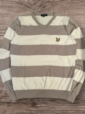 Randig tröja från Lyle & Scott, M - Snygg långärmad tröja från Lyle & Scott i storlek M. Tröjan har breda beige och vita ränder, v-ringad hals och det klassiska gula fågelmärket broderat på bröstet. Mjuk bomullskvalitet och ribbade muddar vid ärmslut och nederkant.