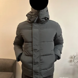 Mörkgrå pufferjacka från Canada Goose - Helt ny grå dunjacka från Canada Goose med huva och dragkedja. Originalpåse och taggar medföljer.