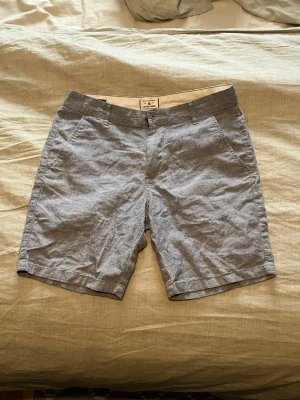 Grå linneshorts från Jack & Jones - Snygga grå shorts från Jack & Jones i mjukt linne. Klassisk passform med midwaist, fickor fram och bak samt dragkedja och knappstängning. Perfekta för varma dagar och chill häng. Diskret logga bak på ena fickan.