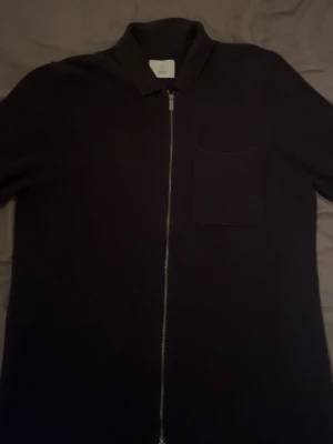 Svart kofta med dragkedja från Bläck storlek S - Stilren svart kofta från Bläck i storlek S. Koftan har en hel dragkedja framtill, klassisk krage och en bröstficka. Perfekt för dig som gillar minimalistisk design och vill ha något enkelt att slänga på sig. Materialet känns mjukt och bekvämt.