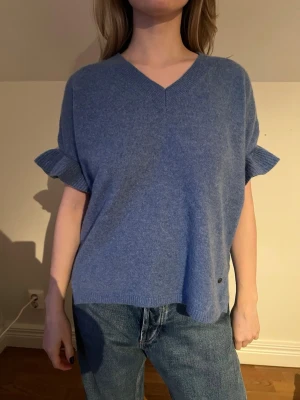 Blå Kashmir tröja med volangärm - Mysig blå Kashmir tröja med v-ringad hals och korta ärmar med volangdetalj. Tröjan har en loose passform och ribbade kanter vid ärmslut och nederkant. Perfekt att styla med jeans för en chill look.