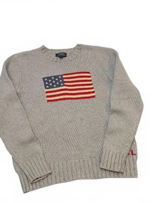 Grå stickad tröja Polo Ralph Lauren - Säljer en grå stickad tröja från Polo Ralph Lauren med amerikansk flagga broderad på bröstet. Tröjan har ribbade muddar vid ärmslut och nederkant samt rund hals. Perfekt för dig som gillar klassisk college-stil.