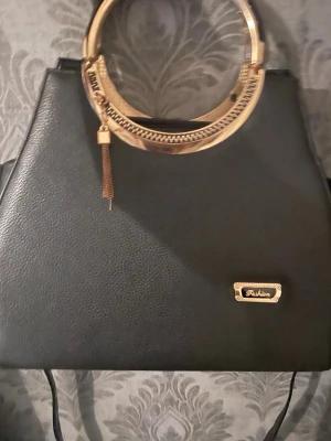 Svart handväska med guldfärgade detaljer - Snygg svart handväska med guldfärgade runda handtag och dekorativ tofs. Väskan har ett stilrent utseende och insidan är fodrad med mönstrat tyg och flera fack med dragkedja. Perfekt för dig som gillar att sticka ut med coola detaljer.